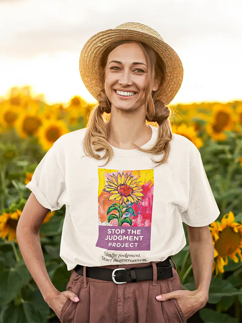 A young smiling woman standing in a sunflower field wearing a white t shirt with the Stop The Judgment Project logo on the front